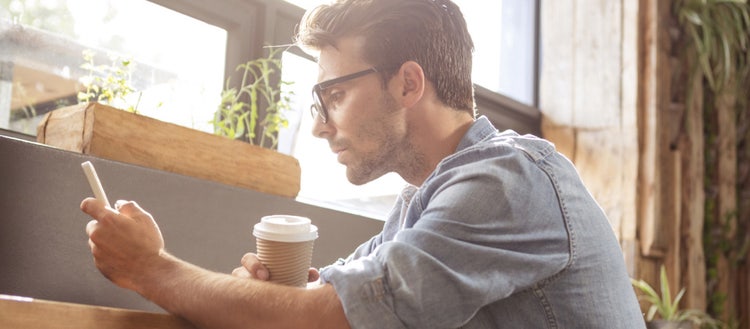 A man reads something on a smartphone while holding a paper coffee cup.