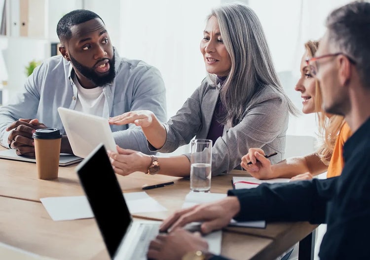 Four people talk while sitting at a table. One woman is holding a tablet. Work supplies and a laptop computer are on the table.