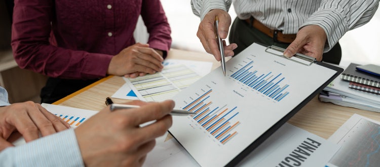 A close-up of a chart being held on a clipboard with three people looking. Their faces are not in the frame. Two people's hands hold pens pointed at the chart.