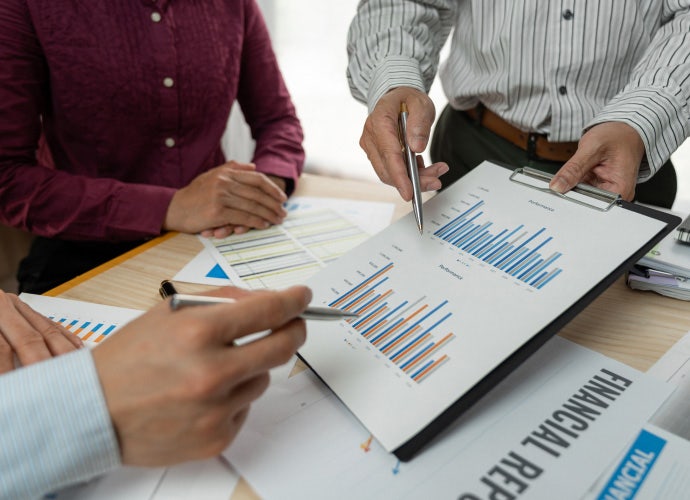 A close-up of a chart being held on a clipboard with three people looking. Their faces are not in the frame. Two people's hands hold pens pointed at the chart.